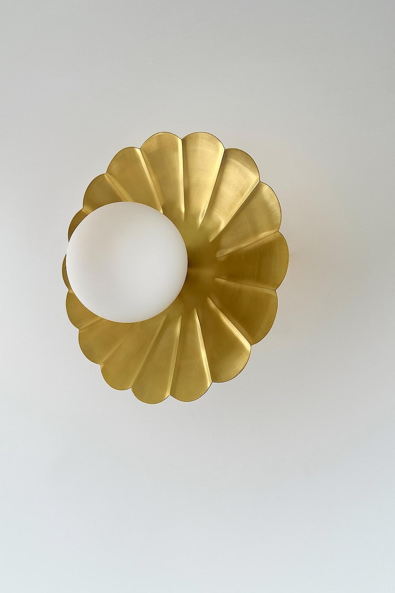 May include: A gold-colored, scalloped wall sconce with a white, spherical light fixture. The sconce has a petal-like design, and the light is positioned in the center. The fixture is mounted on a white wall.