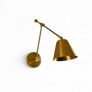 Puede incluir: Aplique de pared de latón con pantalla cónica y brazo ajustable. El aplique tiene una base circular y un acabado metálico cálido. El diseño es minimalista y moderno, adecuado para la iluminación interior.