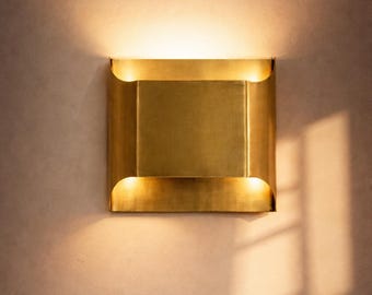 May include: A square, gold-colored wall sconce with a rectangular center and curved edges. The sconce emits a warm light from the top and bottom, casting shadows on the wall. The fixture is mounted on a neutral-toned wall.