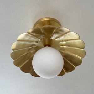 Può includere: Un lampadario da soffitto dorato a forma di conchiglia, con una lampadina a globo bianca al centro. Il lampadario è montato su un soffitto bianco. Il design è moderno e decorativo.