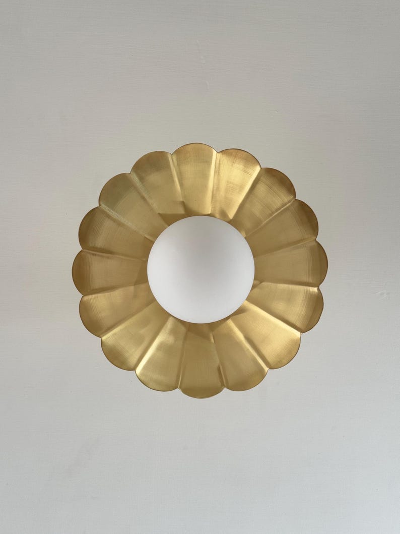 May include: A gold-colored, scalloped-edge ceiling light fixture with a white, spherical light bulb in the center. The fixture has a flower-like design and is mounted on a white ceiling.