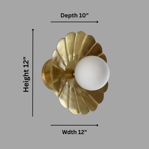 May include: A brass wall sconce with a scalloped design and a white globe light. The sconce is 12 inches wide, 12 inches high, and 10 inches deep. The fixture has a modern, elegant design.