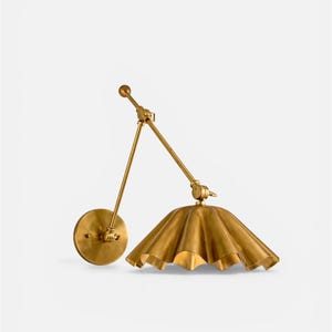 Op de afbeelding: Een messing wandlamp met een verstelbare arm en een geplooide kap. De lamp heeft een ronde wandplaat en een kogelvormig detail aan het einde van de arm. De kap heeft een geschulpte rand.