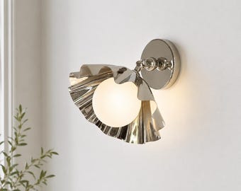 Könnte beinhalten: Eine Wandleuchte mit einem polierten, silberfarbenen Metallsockel und einem passenden, wellenförmigen Schirm in Muschelform. Eine kugelförmige, mattweiße Glaslampe befindet sich in der Mitte des Metallschirms und strahlt ein warmes Licht aus.
