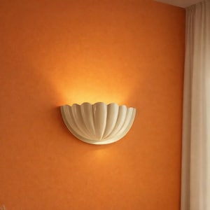 May include: A white, scalloped wall sconce is mounted on an orange wall. The sconce has a curved, petal-like design, and the light shines downward, creating a warm glow. The wall is painted a solid orange color, and a white curtain is visible on the right side.