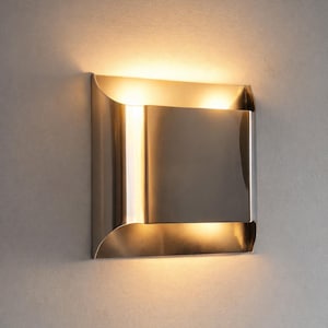 May include: A modern, brushed metal wall sconce with a square design. The sconce has a rectangular center and curved edges, emitting a warm, diffused light. The fixture is mounted on a light gray wall.