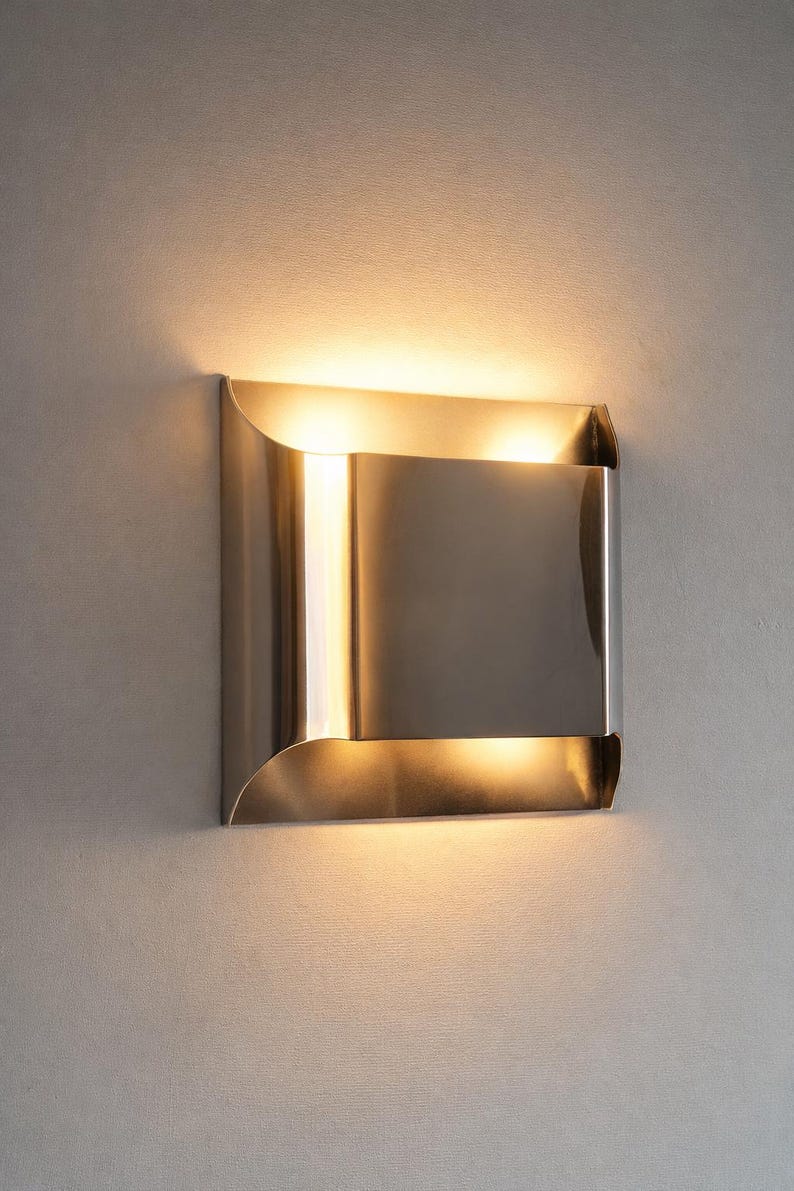 May include: A modern, brushed metal wall sconce with a square design. The sconce has a rectangular center and curved edges, emitting a warm, diffused light. The fixture is mounted on a light gray wall.