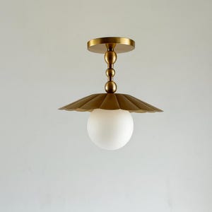 Puede incluir: Una lámpara de techo de color latón con una pantalla festoneada y un globo esmerilado. La lámpara tiene una base redonda y una serie de esferas decorativas que conectan la base con la pantalla. La luz está suspendida sobre un fondo blanco liso.