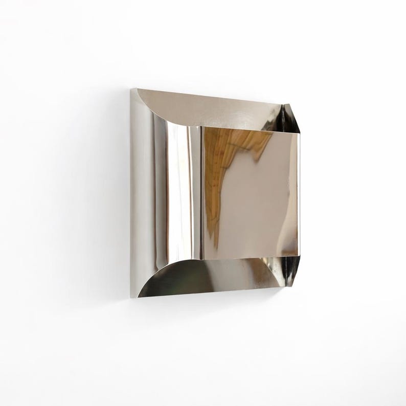 May include: A polished, stainless steel wall-mounted mailbox with a curved top and bottom. The mailbox has a rectangular body with a reflective surface, mounted on a white wall. The design is modern and minimalist.
