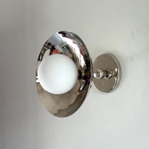 May include: A contemporary wall sconce with a shiny, silver finish. It has an oval shade with a white globe light inside. The sconce is mounted on a circular base with decorative details, suitable for interior lighting.