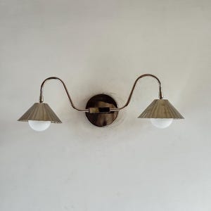May include: A double-arm wall sconce featuring a polished metallic finish. The fixture has a central circular mounting plate with two curved arms extending outward, each supporting a fluted conical shade and a round white glass bulb.