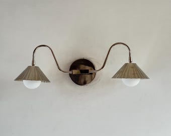 May include: A double-arm wall sconce featuring a polished metallic finish. The fixture has a central circular mounting plate with two curved arms extending outward, each supporting a fluted conical shade and a round white glass bulb.