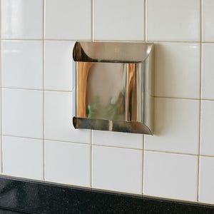May include: A stainless steel, square-shaped wall-mounted dispenser with curved edges. The dispenser is mounted on a white tiled wall above a black countertop. The dispenser reflects the surrounding environment.
