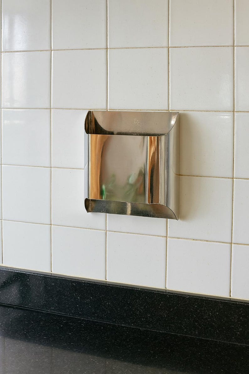 May include: A stainless steel, square-shaped wall-mounted dispenser with curved edges. The dispenser is mounted on a white tiled wall above a black countertop. The dispenser reflects the surrounding environment.