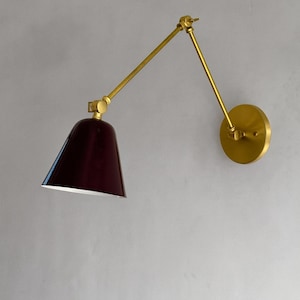 May include: A wall-mounted lamp with a burgundy shade and a brass-colored adjustable arm. The lamp has a circular base and a conical shade, ideal for a bedroom or living room.