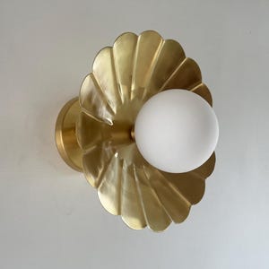 May include: A brass wall sconce featuring a scalloped, shell-like design. The fixture has a round, frosted white globe light in the center. The sconce is mounted on a circular base, creating a decorative lighting element.