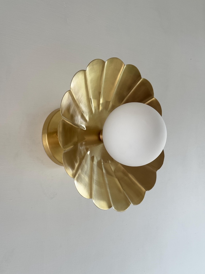 May include: A brass wall sconce featuring a scalloped, shell-like design. The fixture has a round, frosted white globe light in the center. The sconce is mounted on a circular base, creating a decorative lighting element.