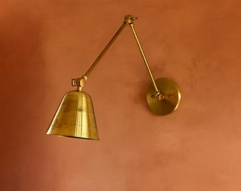 May include: A brass wall lamp featuring a circular mounting plate, an adjustable articulated arm with two segments, and a conical shade. The metal has a polished finish, and the lamp is positioned to extend outward from the wall surface.