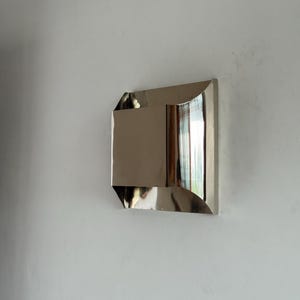 May include: A modern, geometric wall mirror with a polished, metallic finish. The mirror has a rectangular shape with angled and curved edges, reflecting the surrounding environment. It is mounted on a white wall, creating a minimalist aesthetic.