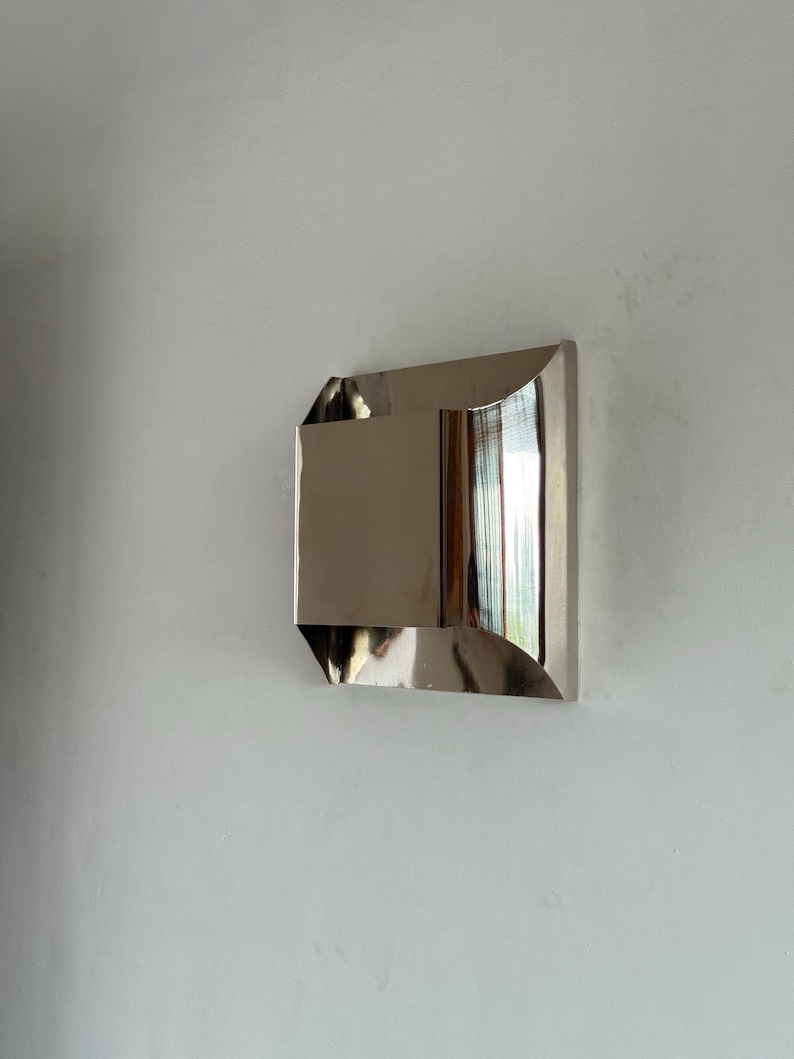 May include: A modern, geometric wall mirror with a polished, metallic finish. The mirror has a rectangular shape with angled and curved edges, reflecting the surrounding environment. It is mounted on a white wall, creating a minimalist aesthetic.