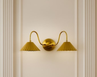 May include: A gold-toned wall sconce with two lampshades. The lampshades are a muted yellow and have a pleated design. The sconce is mounted on a white wall with decorative molding and columns.