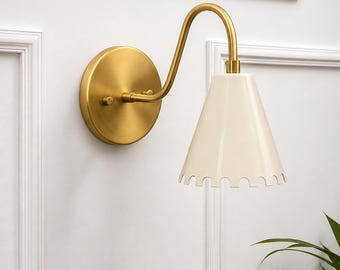 May include: A wall-mounted light fixture featuring a circular brushed brass backplate and a curved brass arm. The lamp includes a conical, off-white shade with a scalloped bottom edge, providing a downward-facing light source for interior spaces.