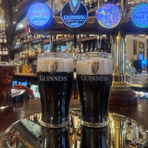 May include: Two glasses of Guinness stout with a creamy head, displayed on a silver tray. The Guinness logo is visible on the glasses. Beer taps with illuminated logos are in the background, including Peroni and Guinness.