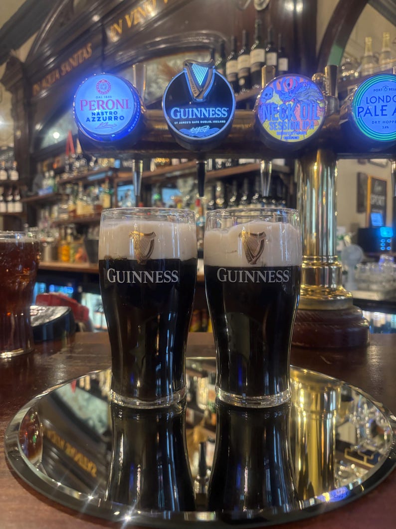 May include: Two glasses of Guinness stout with a creamy head, displayed on a silver tray. The Guinness logo is visible on the glasses. Beer taps with illuminated logos are in the background, including Peroni and Guinness.