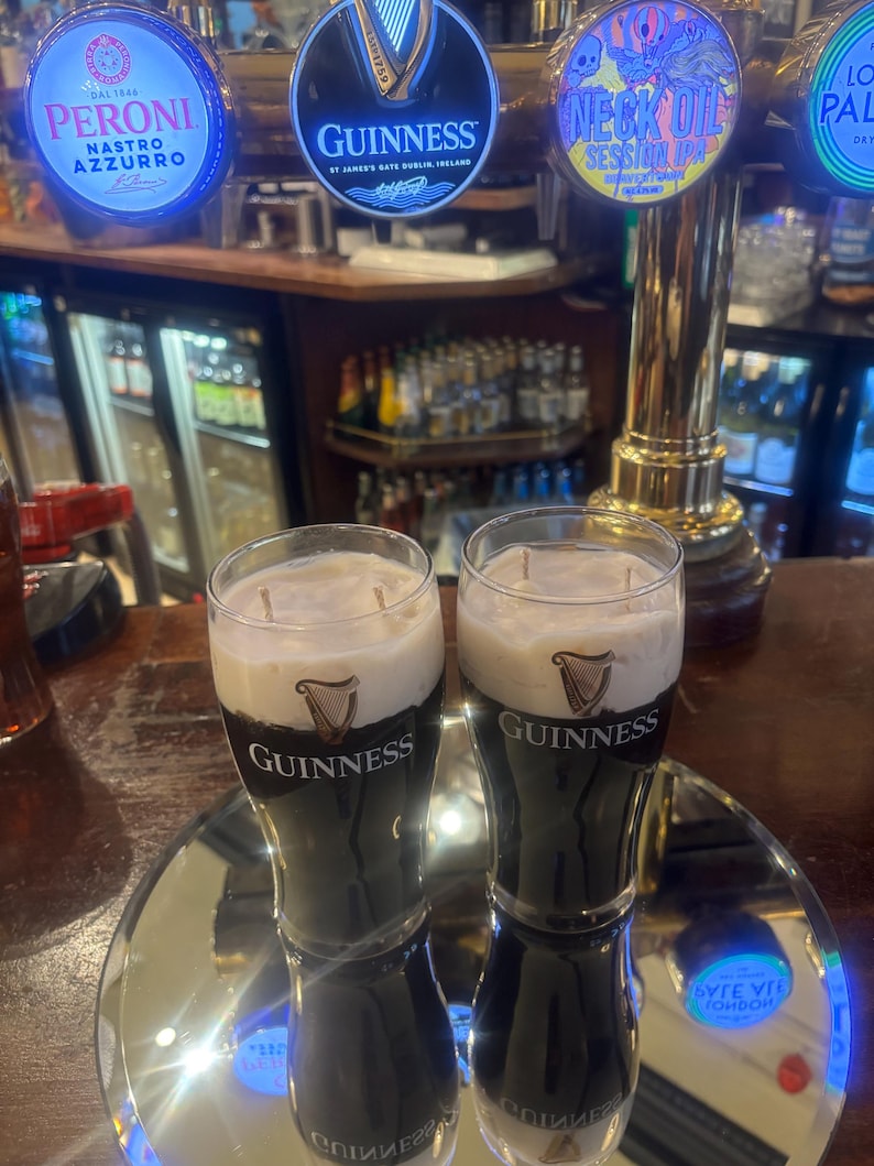 May include: Two glasses of Guinness stout with a creamy head, displayed on a mirrored surface. The Guinness logo is visible on the glasses. Beer taps with illuminated logos for Peroni, Guinness, and Neck Oil are in the background.