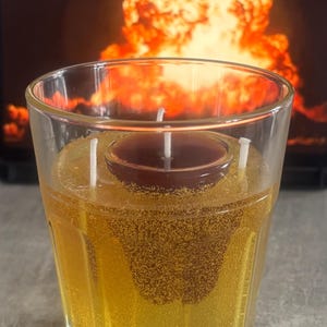 Könnte beinhalten: Eine klare Glaskerze mit einer dunkelbraunen Kerze in der Mitte, umgeben von gelb-goldenem Wachs. Die Kerze hat drei weiße Dochte. Der Hintergrund zeigt eine orange und gelbe Explosion.