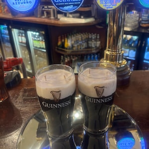 May include: Two glasses of Guinness stout with a creamy head, displayed on a mirrored surface. The Guinness logo is visible on the glasses. Beer taps with illuminated logos for Peroni, Guinness, and Neck Oil are in the background.