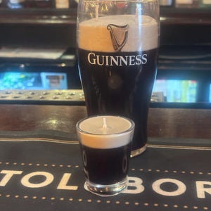May include: Two Guinness-themed candles, a pint and a shot glass, mimicking the appearance of the Irish stout. The candles are dark brown with a creamy white top, and the Guinness logo is visible on the larger candle.