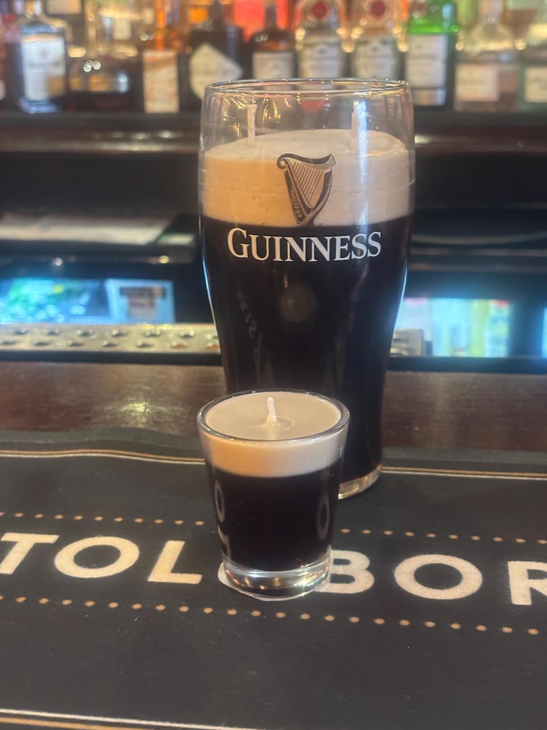 May include: Two Guinness-themed candles, a pint and a shot glass, mimicking the appearance of the Irish stout. The candles are dark brown with a creamy white top, and the Guinness logo is visible on the larger candle.