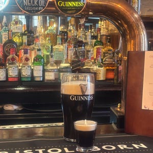 May include: A pint of Guinness and a shot of the same stout, both with a creamy head, on a bar. The Guinness logo is visible on the pint glass. The bar has a black surface with the words "BRISTOL BORN" printed on it. A selection of bottles are in the background.