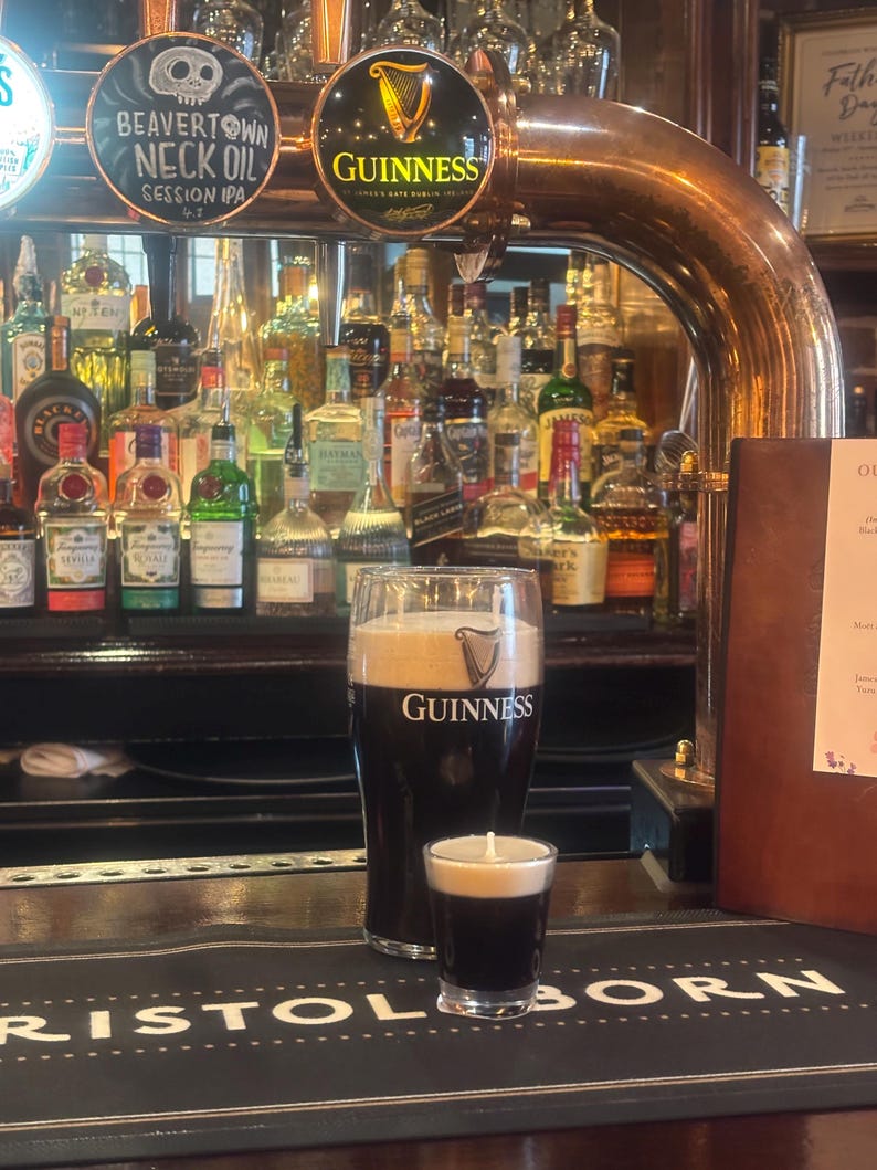 May include: A pint of Guinness and a shot of the same stout, both with a creamy head, on a bar. The Guinness logo is visible on the pint glass. The bar has a black surface with the words "BRISTOL BORN" printed on it. A selection of bottles are in the background.
