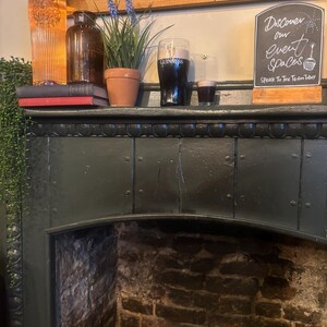 May include: Interior shot of a pub fireplace with a dark green mantelpiece. On the mantel are a bottle, a brown glass jar, a potted plant, a pint of Guinness, and a small glass of stout. A chalkboard sign reads "Discover our event spaces."