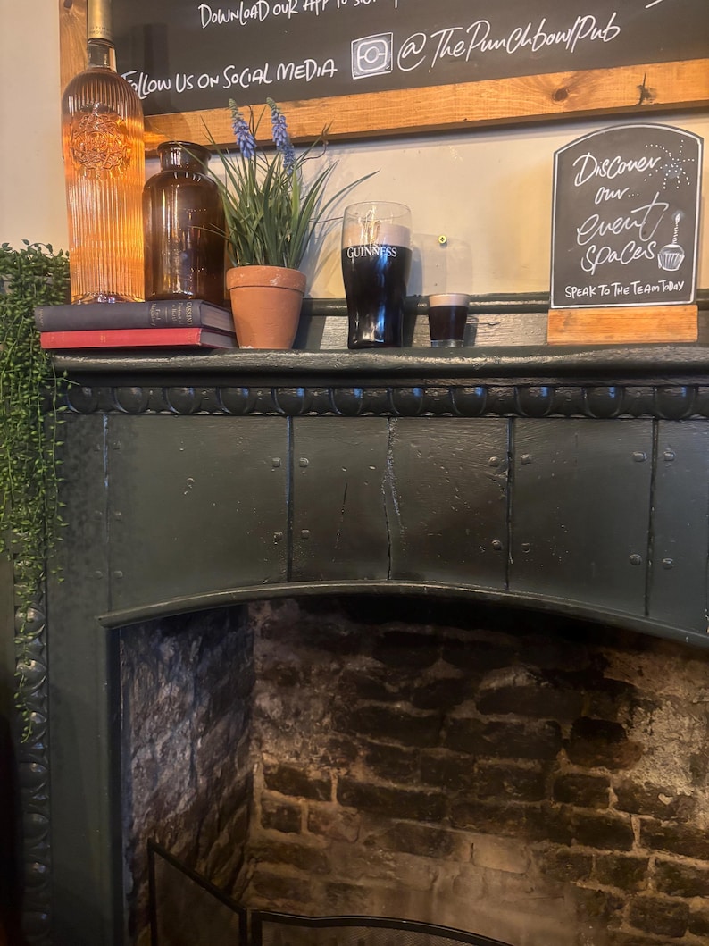 May include: Interior shot of a pub fireplace with a dark green mantelpiece. On the mantel are a bottle, a brown glass jar, a potted plant, a pint of Guinness, and a small glass of stout. A chalkboard sign reads "Discover our event spaces."