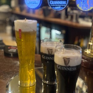 May include: A candle shaped like a tall glass of beer with a foamy head, and two glasses of Guinness stout. The candles sit on a mirrored surface, with beer taps in the background.
