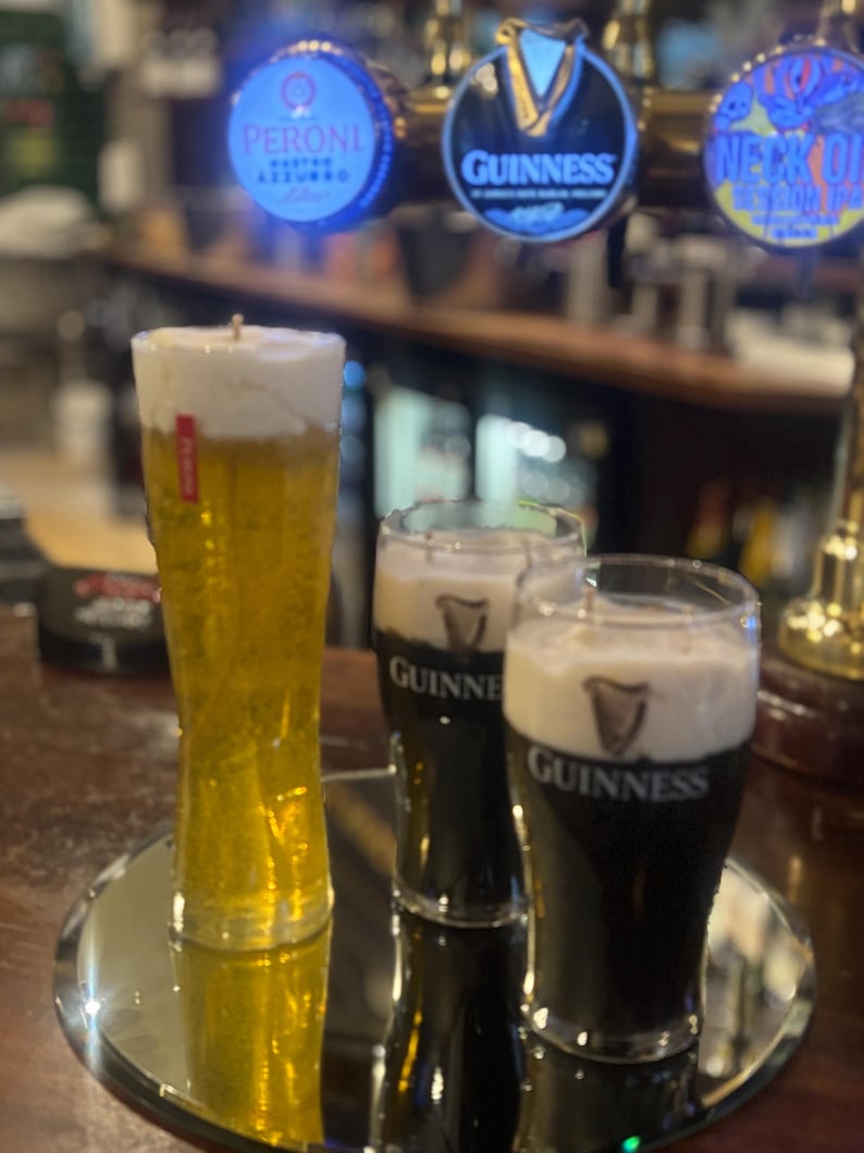 May include: A candle shaped like a tall glass of beer with a foamy head, and two glasses of Guinness stout. The candles sit on a mirrored surface, with beer taps in the background.