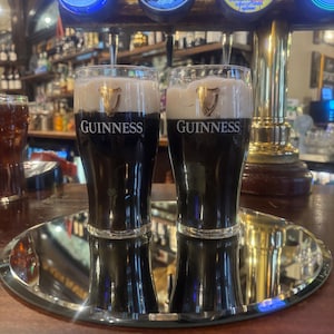 May include: Two glasses of Guinness stout on a mirrored tray, with the Guinness logo prominently displayed. The dark beer is topped with a creamy white head. The background shows beer taps and a bar setting.