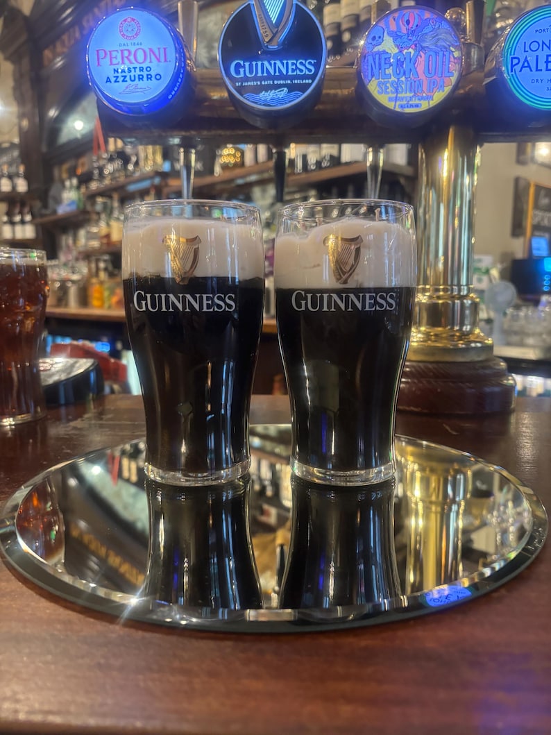 May include: Two glasses of Guinness stout on a mirrored tray, with the Guinness logo prominently displayed. The dark beer is topped with a creamy white head. The background shows beer taps and a bar setting.