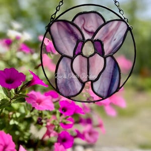 May include: A stained glass floral suncatcher with a black metal frame and chain. The flower design features shades of purple and pink glass. The text "Luna Glass Garden" is visible on the bottom left of the suncatcher.