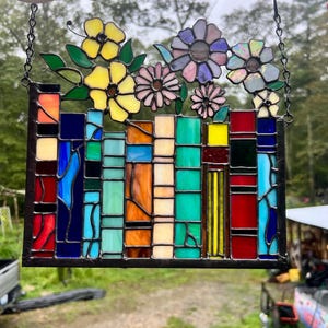 Op de afbeelding: Een glas-in-loodraam met een kleurrijk bloemmotief. Het paneel heeft een rechthoekige basis met diverse gekleurde glazen blokken. Boven de blokken bevinden zich bloemen in gele, roze en paarse tinten, met groene bladeren. Het paneel wordt opgehangen aan een ketting.