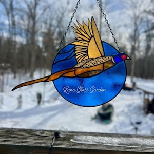 May include: Stained glass art piece featuring a pheasant in flight, set against a blue circular background. The bird has gold, brown, and blue hues, with a detailed wing design. The piece is suspended by a chain and has the text "Luna Glass Garden".