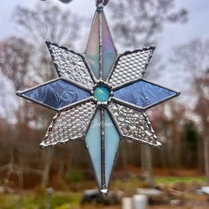 May include: A handcrafted stained glass star ornament. The star features various shades of blue, white, and iridescent glass, with textured and patterned sections. A small, round, turquoise-colored glass piece is at the center. The text "Luna Glass Garden" is visible.