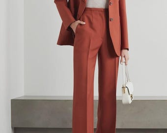 May include: A rust-colored pantsuit, featuring a blazer with a two-button closure and wide-leg trousers. The outfit is paired with a light-colored top and white sandals. A small white handbag with a gold-tone clasp completes the ensemble.
