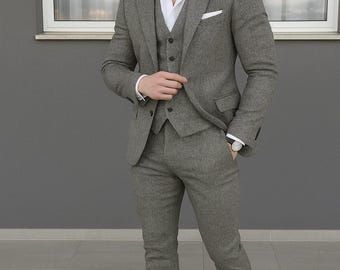 May include: A three-piece grey suit with a white dress shirt and black loafers. The suit includes a blazer, waistcoat, and tailored trousers. The man is wearing a watch. The suit is made of a textured fabric.
