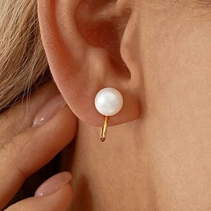 May include: A close-up of a gold-colored earring with a white pearl. The earring is a small, delicate design, with the pearl positioned at the earlobe. The earring is a simple, elegant piece of jewelry.