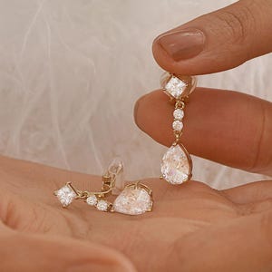 May include: A pair of gold-toned drop earrings featuring a clear pear-shaped stone at the base, suspended from a vertical arrangement of two smaller round stones and a square-cut stone at the top, all set in a polished metallic frame.