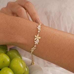May include: A gold-toned chain bracelet featuring a series of delicate flower-shaped charms adorned with small, clear, sparkling stones. The bracelet has a thin, adjustable chain design that rests against the wrist, showcasing a floral motif.